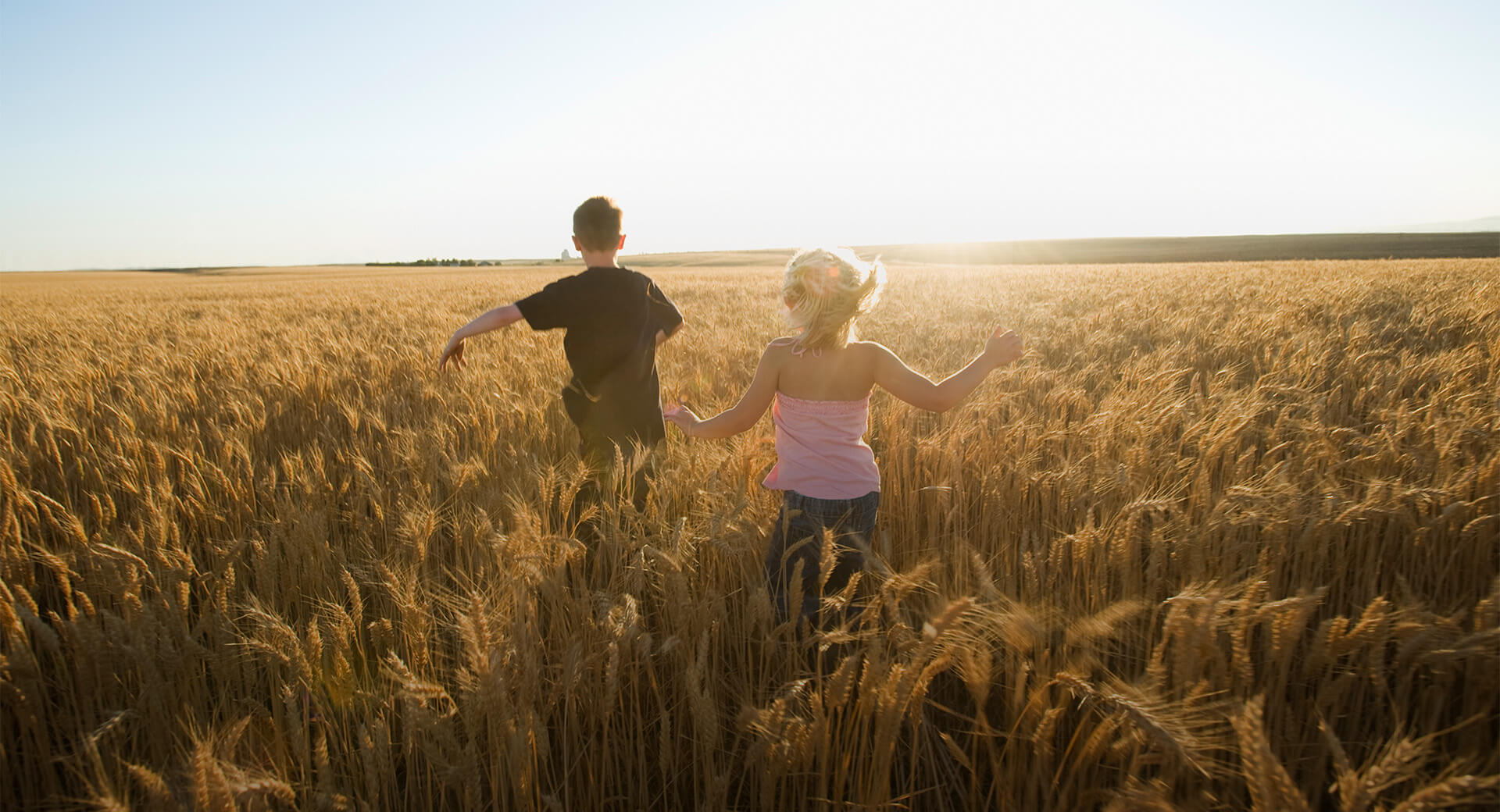 Two children running through a wheat field with the sun setting in the background